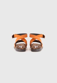 Oransje semskede sandaler med kryssende stropper, åpen bak, og en leopardmønstret innersåle. Flat såle med en brun gummibase.