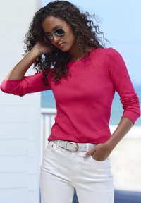 Rotes langärmliges, eng anliegendes Hemd aus strukturiertem Stoff, kombiniert mit weißen Jeans und einem hellgrauen Gürtel mit goldener Schnalle. Modell trägt eine Sonnenbrille.