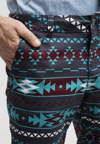 Ruka v kapse, na sobě tmavé šortky s geometrickými kmenovými vzory v tyrkysové, vínové a bílé barvě a modrou riflovou košili.