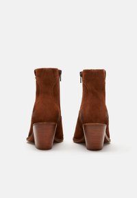 Botines de ante marrón con tacón apilado, que cuentan con costuras decorativas y un cierre de cremallera en el lateral. Textura suave con un diseño clásico.