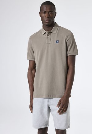 Lichtbruin poloshirt met een kraag, korte mouwen en een blauw logopatch op de borst; gecombineerd met lichte denim shorts.