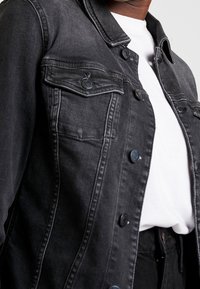 Czarna kurtka dżinsowa z dwoma kieszeniami na piersi, ciemnymi guzikami i widocznymi przeszyciami. Noszona na białej koszulce, prezentująca casualowy design.