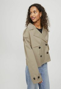 Giacca doppiopetto accorciata in tessuto beige, con grandi bottoni neri, ampi rever e una vestibilità rilassata. Indossata sopra un top nero.