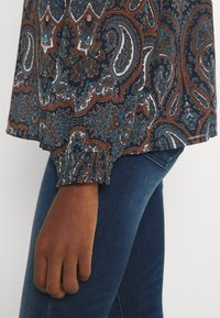 T-shirt à manches longues avec un motif cachemire en bleu marine, orange et turquoise. Présente des poignets froncés. Porté avec un jean en denim bleu.