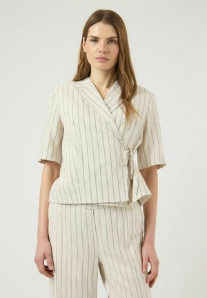 Kvinne med lys brunt hår har på seg en beige kortermet innpakket blazer med tynne striper og matchende bukser med tynne striper, og står foran en ensfarget bakgrunn.