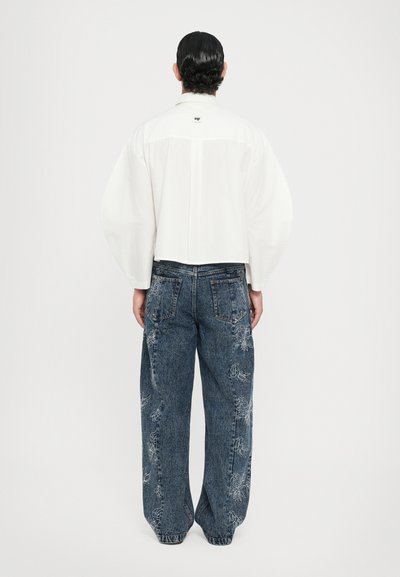 Camisa blanca, recortada, de manga larga con un detalle de pliegues en la parte posterior, combinada con vaqueros de mezclilla anchos y azules con bordados florales.
