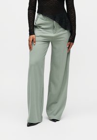 Moteris dėvi laisvus šviesiai žalius plačius kelnes, juodus smailius batus ir juodą ilgomis rankovėmis nėriniuotą viršutinę dalį su asimetrišku apačios kraštu.