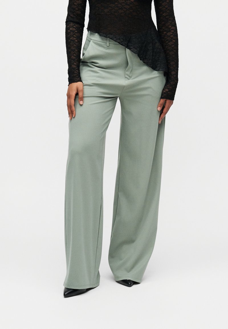 Moteris dėvi laisvus šviesiai žalius plačius kelnes, juodus smailius batus ir juodą ilgomis rankovėmis nėriniuotą viršutinę dalį su asimetrišku apačios kraštu.