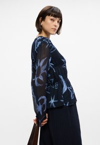 Blouse noire transparente avec des motifs abstraits bleus, manches longues, taille froncée, assortie à un pantalon à rayures. Texture lisse.