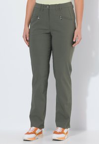 Persona vistiendo pantalones rectos verde oliva con bolsillos delanteros con cremallera y zapatillas naranja y blancas, de pie contra un fondo blanco.