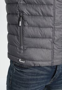 Herrgrå pufferjacka med en quiltad design, som har ett ficka med dragkedja och en Superdry-logotagg nära fållen. Slät, texturerad tyg.