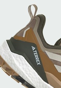 Buty outdoorowe Terrex charakteryzują się brązowo-zieloną, wysokościowo regulowaną cholewką, teksturowaną siateczką, gumową podeszwą oraz białą podeszwą środkową z akcentami logo.
