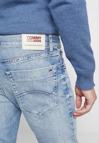 Närbild av ljusblå denimjeans med Tommy Jeans-etikett och liten flagglogotyp på bakfickan, kombinerat med en blå tröja.