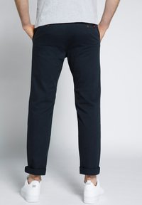 Chino scuri navy con vestibilità slim, orli arrotolati e due tasche posteriori. Realizzati in cotone con una consistenza morbida, abbinati a sneaker bianche.