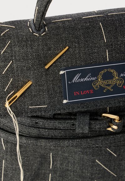 Mala de tecido cinza com detalhes de costura brancos. Apresenta acessórios em ouro e um rótulo azul com o logótipo Moschino e o texto "IN LOVE."