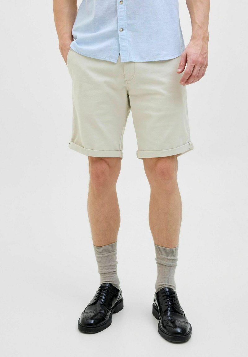 Light beige rolled shorts made of cotton, paired with grey socks and black leather shoes. Features a button-down shirt in light blue.