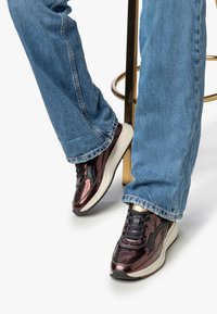 Persona che indossa sneakers metallici color borgogna e jeans blu, seduta con i piedi su un pavimento bianco e un poggiapiedi in metallo dorato dietro di sé.