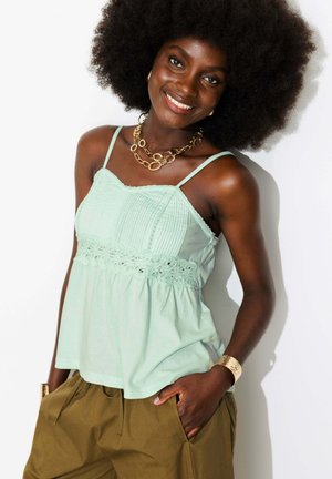 Femme souriante avec un grand afro portant un haut sans manches vert clair, un collier chaîne en or, un bracelet et un pantalon vert olive les mains dans les poches.
