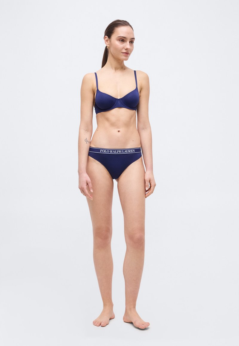 Γυναίκα όρθια, ξυπόλητη, φοράει μπλε σκούρο σουτιέν και εσώρουχα με το λογότυπο Polo Ralph Lauren, μπροστά σε απλό φωτεινό φόντο.