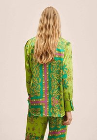 Persoon met lang golvend blond haar, gekleed in een groene satijnen blouse met patroon en bijpassende broek, staat met de rug naar de camera tegen een effen achtergrond.