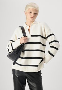 Sweter dziany w kolorze ecru z czarnymi poziomymi paskami, z kołnierzykiem i warkoczowym splotem. Połączony z czarnymi skórzanymi spodniami.