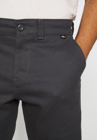 Gros plan d'une personne portant un pantalon gris foncé avec un bouton et une boucle de ceinture, la main reposant dans la poche avant.