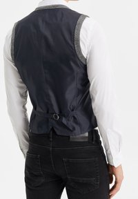 Mörk marinblå väst med texturerade grå axelpartier, justerbar baksidedel, i kombination med en vit skjorta och svarta jeans. Slät tygfinish.