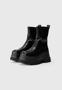 Botas de cuero negras hasta el tobillo con una suela gruesa de plataforma, que presentan una textura suave, lados elásticos y lengüetas para facilitar su uso.