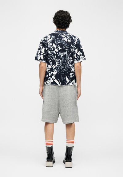 Oseba stoji obrnjena stran, oblečena v črno-belo s cvetličnim in plesnim potiskom majico, sive kratke hlače do kolen, črtaste nogavice in črne škornje.