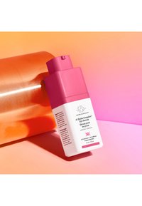Pink bottle of Drunk Elephant A-Shaba Complex Eye Serum with white label and black text, placed on a reflective orange background, angled.
