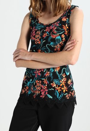 Kobieta w czarnym topie bez rękawów z kolorowym kwiatowym nadrukiem i falistym koronkowym wykończeniem, ze skrzyżowanymi ramionami, w połączeniu z czarnymi spodniami.