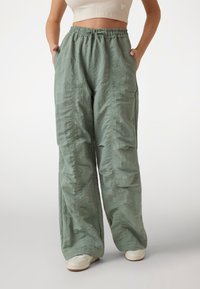 Pantaloni larga de culoare verde deschis, confecționați dintr-un material texturat. Au un elastic în talie și buzunare laterale. Fără modele sau accesorii vizibile.