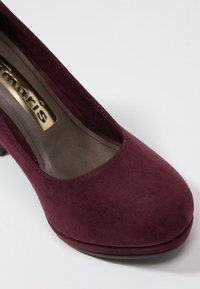 Burgunderfärgad mockaklackskor med rund tå, slät textur och plattformdesign. Guldfärgad märkning synlig på innersulan.