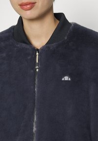 Jachetă bomber din blană sintetică bleumarin, cu guler rib din negru, închidere cu fermoar și un mic logo brodat pe pieptul stâng.