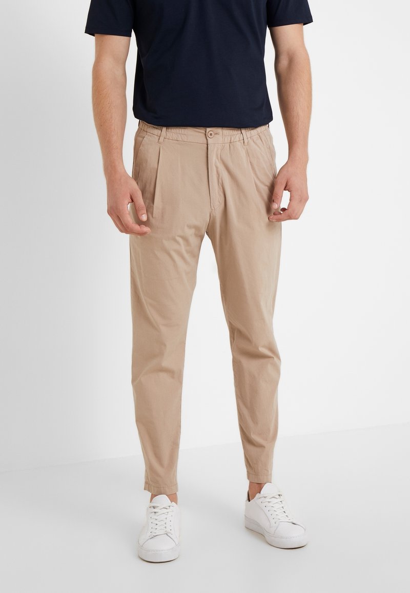 Beżowe spodnie chino z fałdami, o zwężanym kroju, wykonane z gładkiej bawełnianej tkaniny, zapinane na guzik, zestawione z białymi trampkami i granatowym t-shirtem.