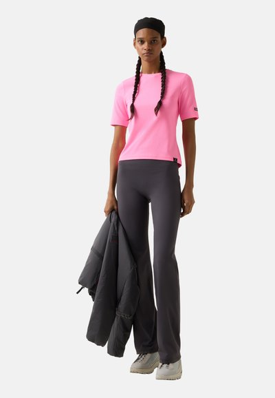 Junge Frau in pinkem kurzärmligem Shirt, dunkelgrauer ausgestellter Hose, weißen Turnschuhen und schwarzem Stirnband, die eine dunkelgraue Jacke in einer Hand hält.