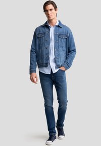 Mann geht, trägt eine blaue Jeansjacke, hellblaues Hemd, blaue Jeans und marineblaue Sneakers vor grauem Hintergrund.