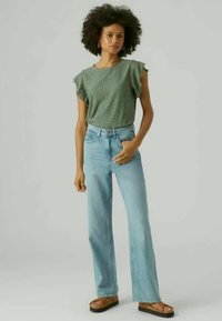 Grøn øjelet top med flæser på ærmerne, parret med lyseblå høj talje jeans og brune platform sandaler. Ensfarvet baggrund.