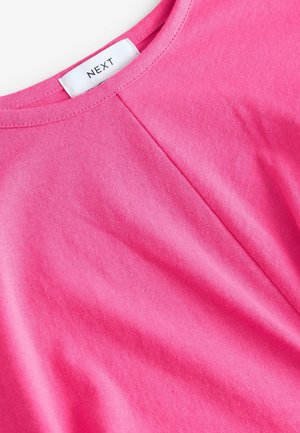 Camiseta de algodón rosa con cuello redondo, sutil detalle de pliegues y una etiqueta de marca en el cuello. Textura suave y tejido ligero.