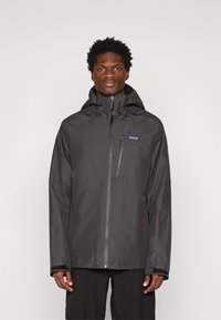 Čierna vodotesná bunda so zipsom vpredu, nastaviteľnou kapucňou a elastickými manžetami. Obsahuje malé logo na hrudi a má štýlový dizajn.