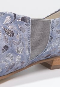 Gros plan sur une chaussure en daim bleu avec des découpes florales argentées, un panneau latéral élastique gris et un petit talon en bois.