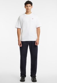 T-shirt bianco in cotone a maniche corte, con un piccolo logo. Abbinato a jeans di denim blu scuro. Modello con scarpe nere.