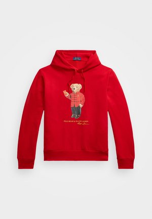 Czerwona bluza z kapturem z niedźwiedziem w czerwonej kurtce i czarnych spodniach, trzymającym dzwonek, z napisem „Polo Bear by Ralph Lauren” poniżej.