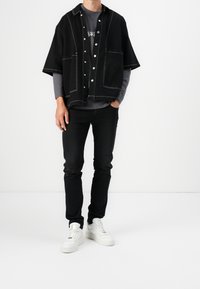 Czarna, oversize kurtka koszulowa z białym przeszyciem, z dużymi kieszeniami, nałożona na szary t-shirt, w zestawie z czarnymi dżinsami i białymi trampkami.