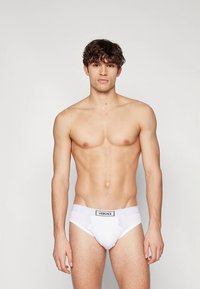 Culotte en coton blanc avec taille élastique, marquée "VERSACE". Présente une coupe classique et une texture lisse. Le mannequin est debout, en position verticale.