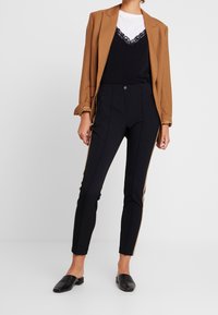 Pantalons noirs ajustés avec poches zippées et rayures latérales beige, associés à un haut noir bordé de dentelle et un blazer marron.