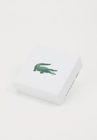 Valge ristkülikukujuline kast tekstuuriga pinnaga, millel on roheline krokodilli logo ja ühel küljel on embosseeritud "LACOSTE".