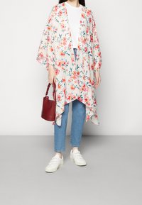 Kimono floral léger en tissu fluide avec des motifs rouges et verts éclatants. Porté avec un jean bleu et des baskets blanches, tenant un sac rouge.
