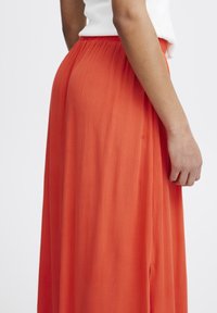 Flödande orange kjol i lätt tyg, med en smockad midja och vertikal textur, som draperar ner till golvet med sidoklyftor.
