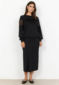 Schwarzes Kleid mit langen Ärmeln, Spitzenakzenten, tailliertem Oberteil und einem Midirock mit einem Schlitz vorne, kombiniert mit schwarzen Ballerinas.